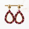 Bordeaux rode druppel oorbellen met fonkelende paarse en rode kralen, goudkleurige ovale oorstekers en open druppelvorm. Elegante sieraden van Seyso.