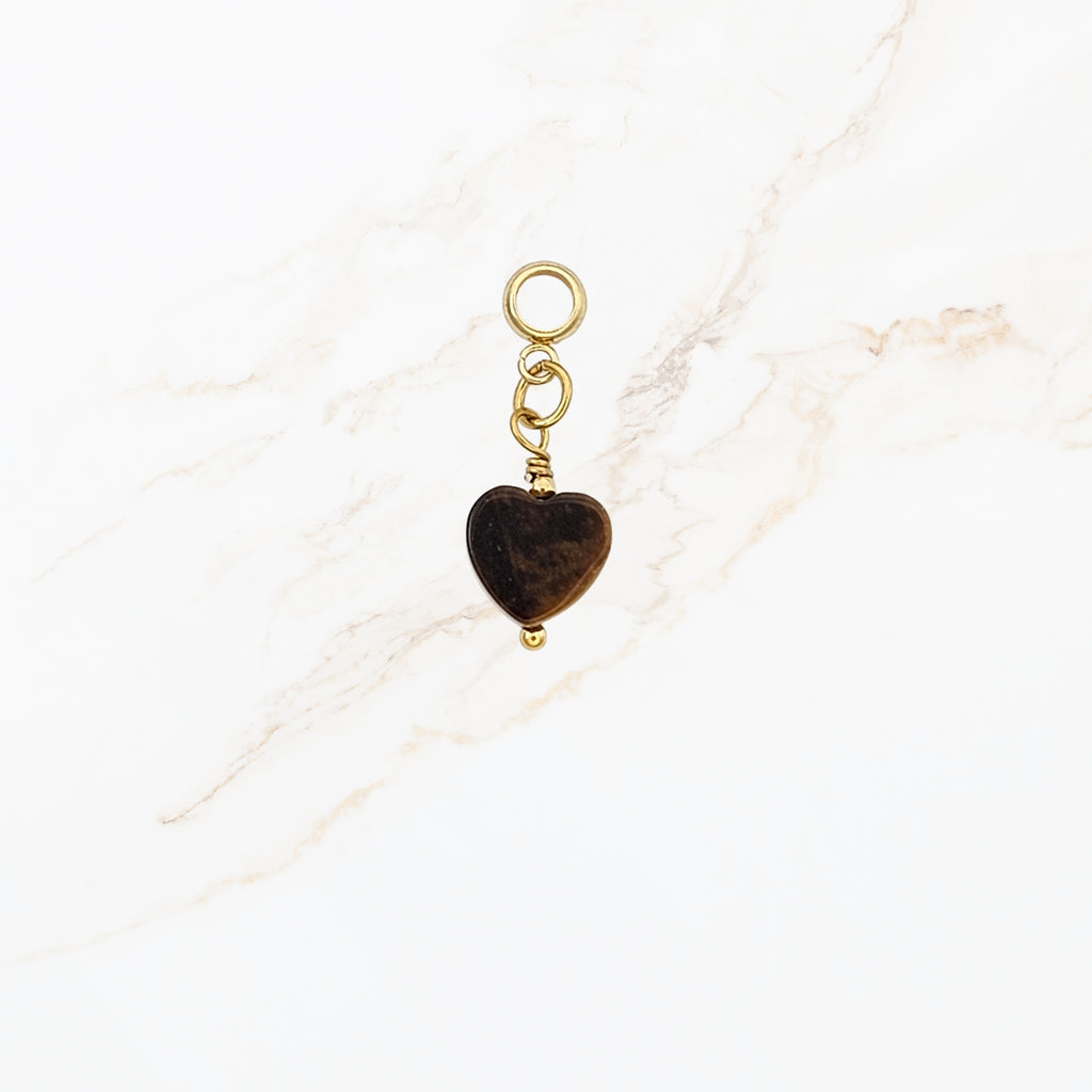 Gouden bedel met een donkerbruin hartje van steen of glas, bevestigd aan een klein schakeltje met een rond oogje, tegen een lichte marmerachtige achtergrond.