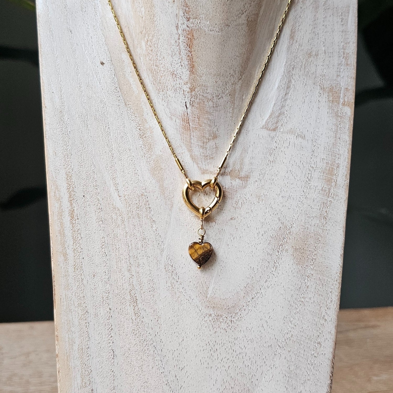 Gouden bedel met een donkerbruin hartje van steen of glas, bevestigd aan een klein schakeltje met een rond oogje, tegen een lichte marmerachtige achtergrond.